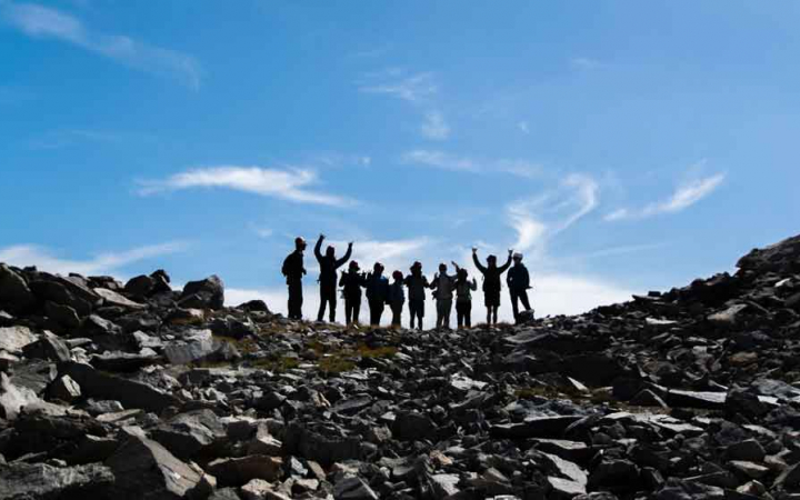 A group of students stand along a rocky ridge under blue skies. A couple of them raise their arms in the air in an apparent gesture of success. 